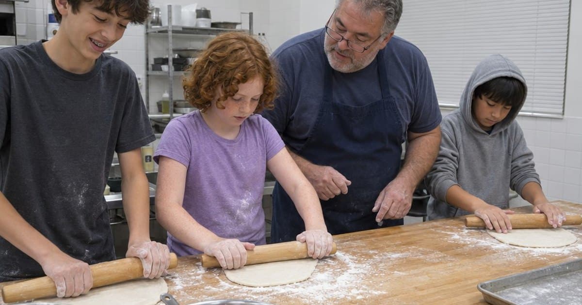 Three tweens roll out dough with wooden rolling pins at a floured wooden counter while their cooking teacher, in glasses and a navy apron, watches and helps in a bright teaching kitchen