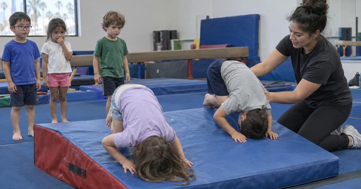 A coach kneels on a blue gym mat helping two young kids do forward rolls while three small children wait their turn in a bright gym