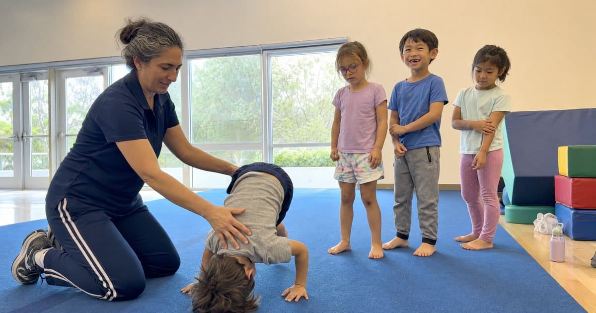 A coach kneels on a blue gym mat helping a small boy do a forward roll while three young kids wait their turn in a bright community gym