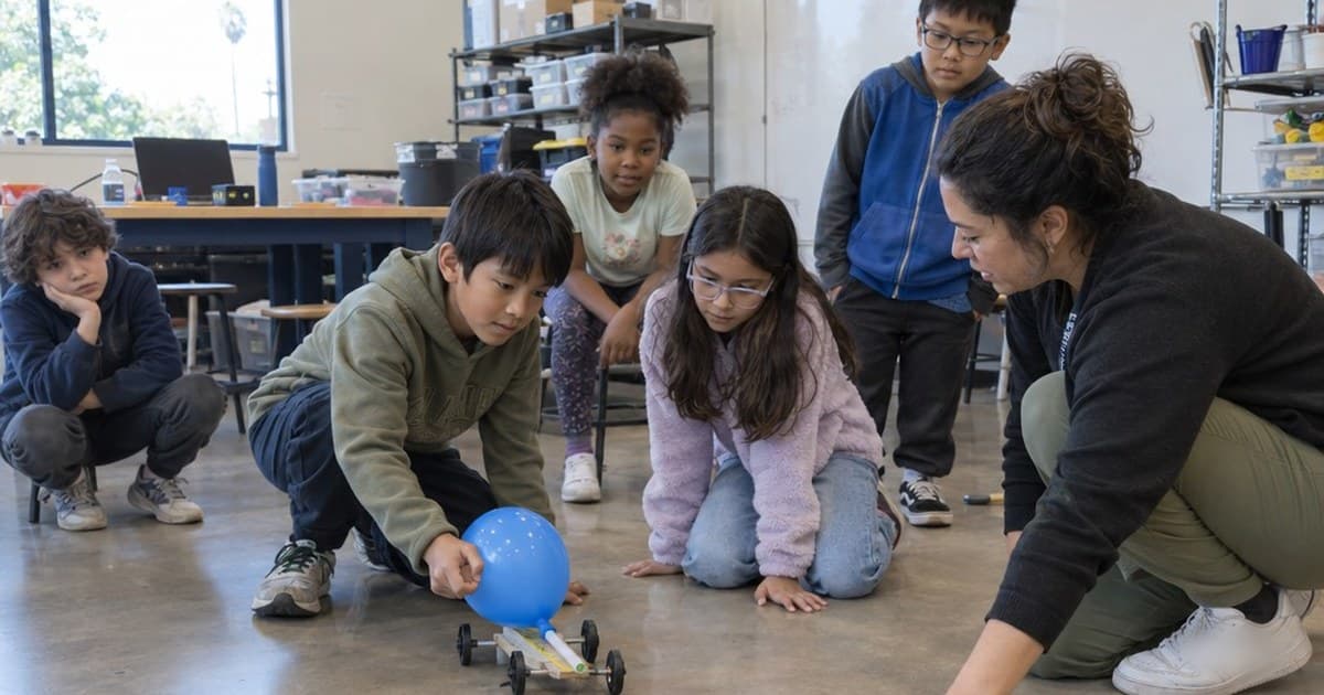 Kids gather on a bright classroom floor around a boy launching a balloon-powered car while their instructor kneels beside them to watch