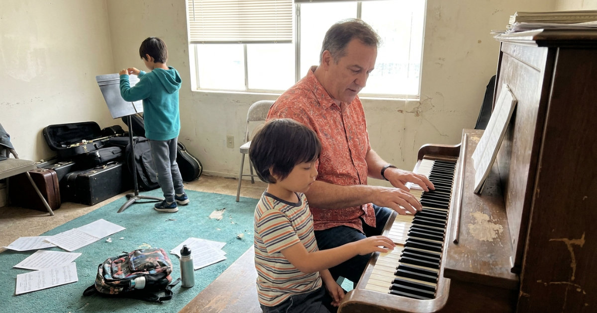 A piano teacher sits beside a young student at an upright piano inside a small studio