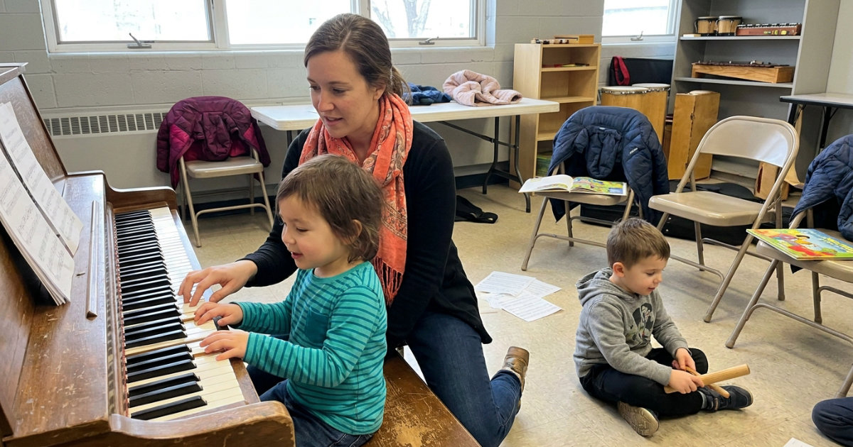 A music teacher kneels beside a small child seated at an upright piano in a bright community center classroom