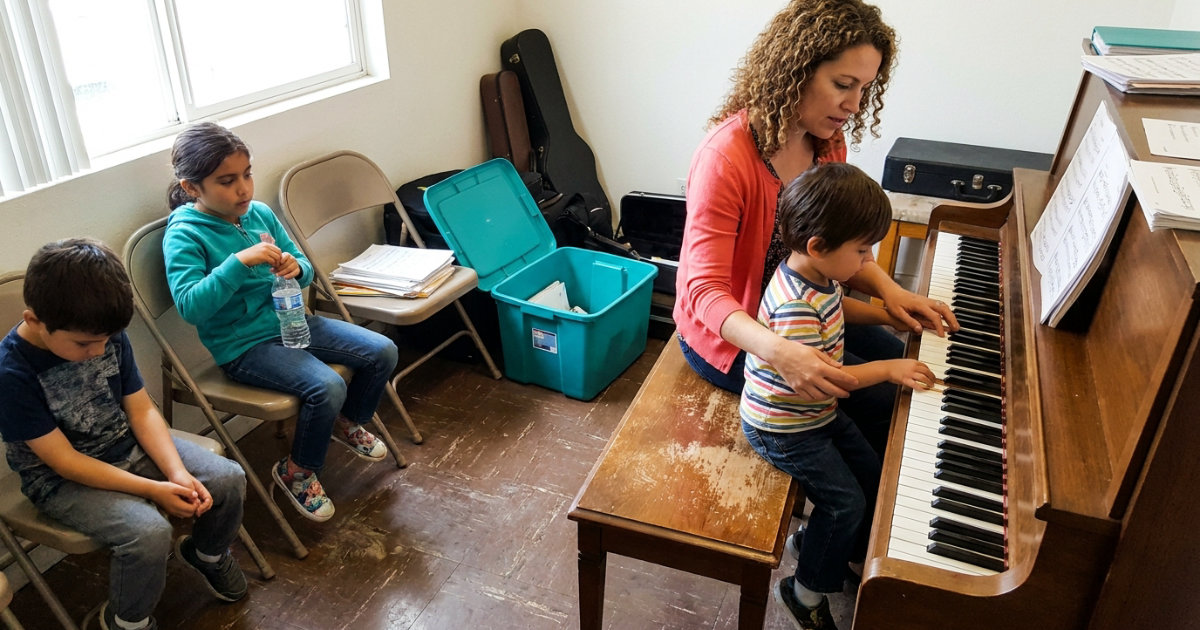 A piano teacher sits beside a young child on a bench in a small studio
